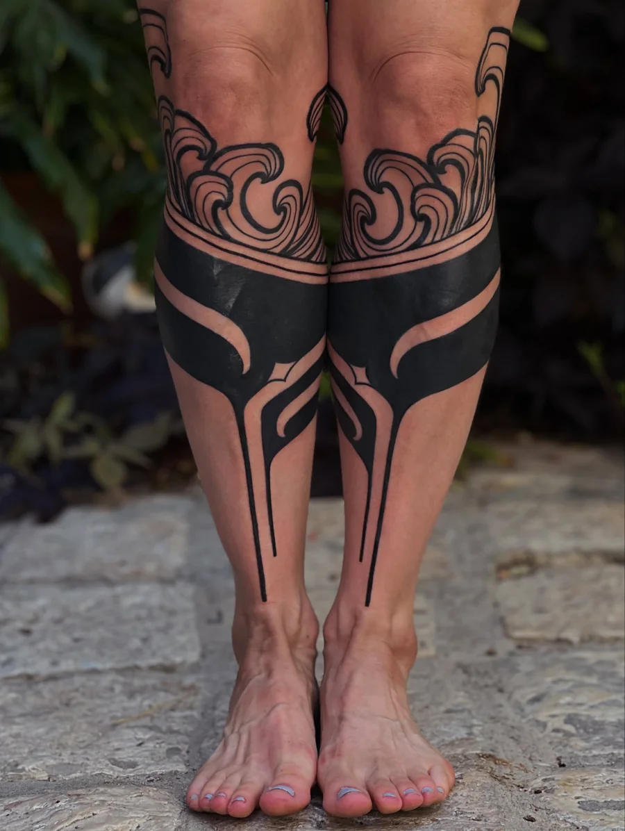 Matching ornamental blackwork tattoos covering both shins with sharp flowing forms and curling linework above.