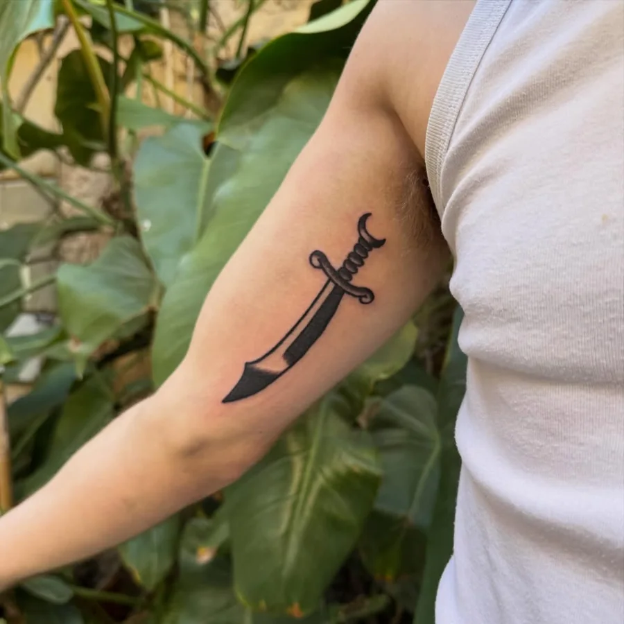 A small curved sword tattoo in bold black linework on the upper arm.