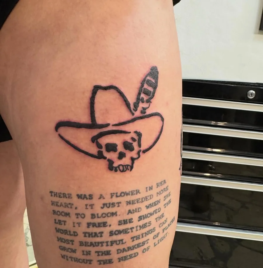 A skull wearing a wide-brimmed cowboy hat with a feather, above an existing text tattoo.