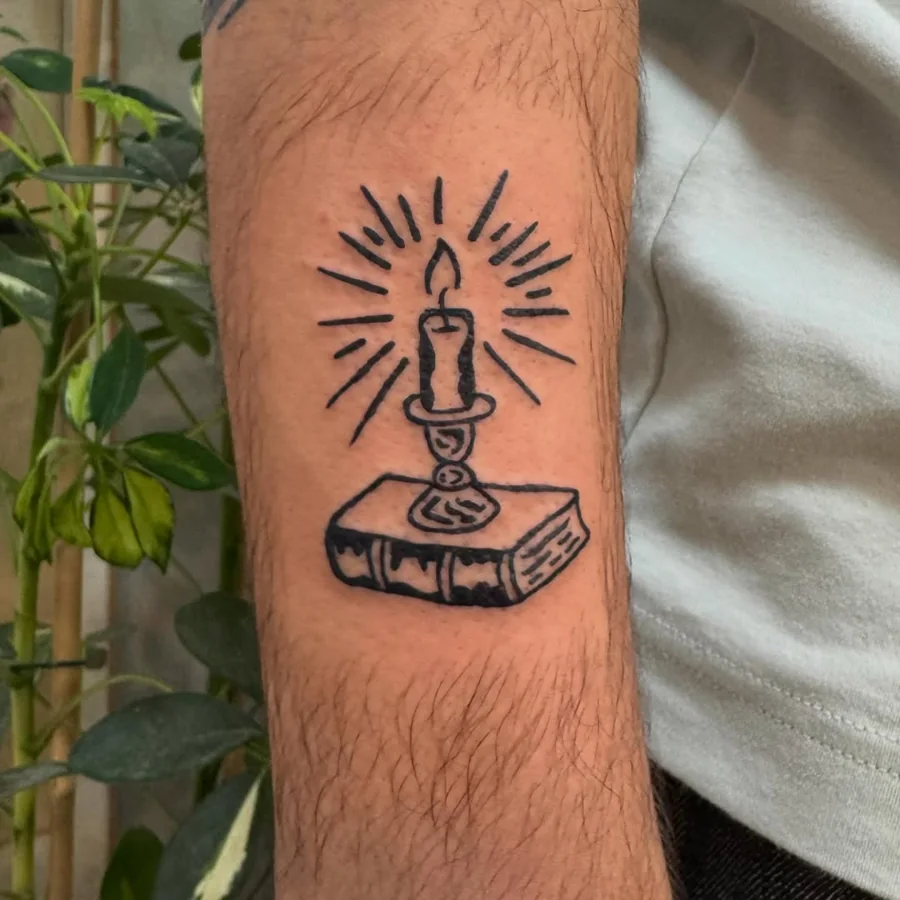 A burning candle with radiating lines on a thick book, drawn in black linework on the forearm.
