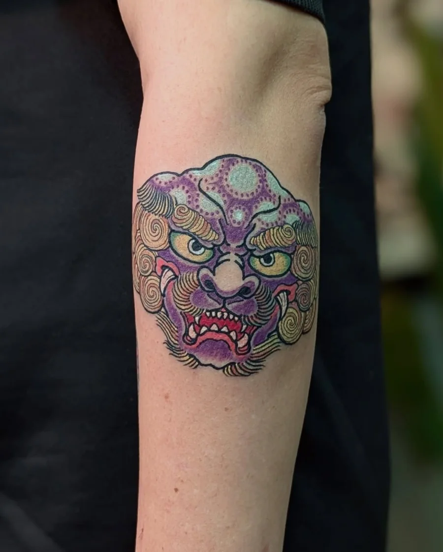 A colorful ornamental guardian face tattoo in purple, green, and gold on the inner forearm.