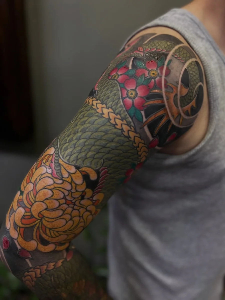 A colorful Japanese-style sleeve with a green scaled serpent, red blossoms, a yellow chrysanthemum, and dark flowing background.