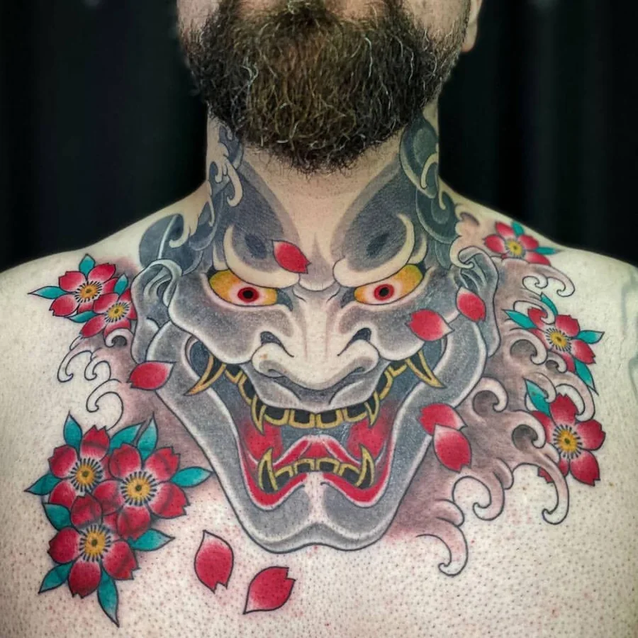A large Japanese hannya mask in grey, red, and yellow across the neck and chest, surrounded by red flowers and swirling clouds.